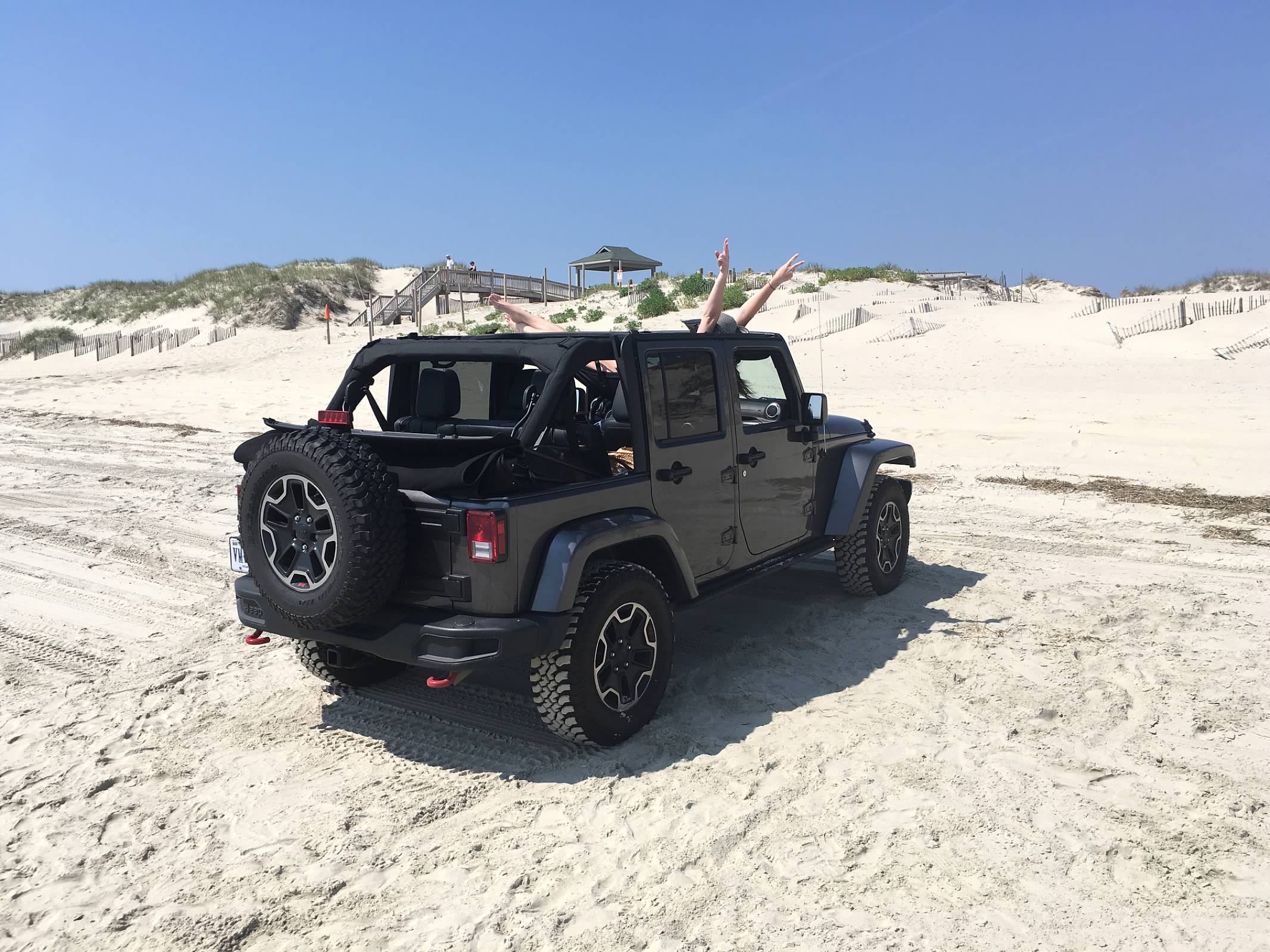A dark gray Jeep Wrangler parked on a sandy beach with dunes and blue sky in the background. A person is sitting inside the vehicle with both arms raised in the air.